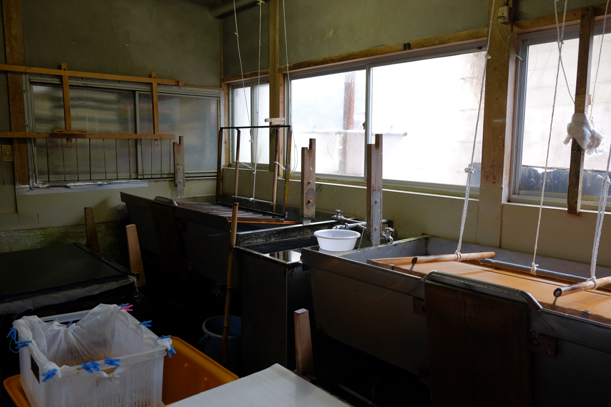 Intérieur d'un atelier avec plusieurs bacs de fabrication de papier, cadres de séchage et panier en plastique, tons verts et bruns tamisés
