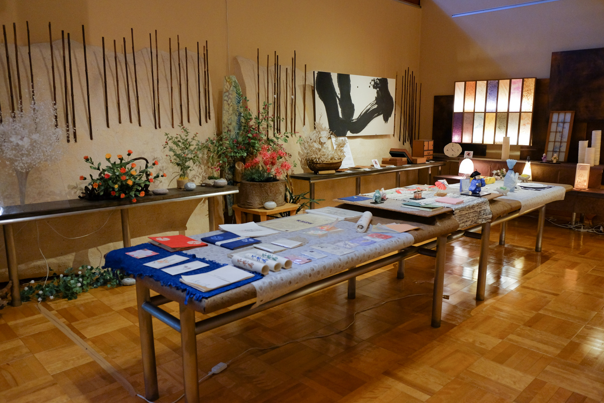 Salle d'exposition avec de longues tables présentant divers objets en papier artisanal, décorations et œuvres murales, ambiance chaleureuse bois et blanc