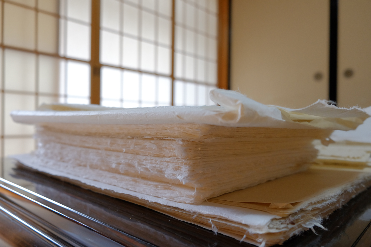 Gros plan de feuilles de papier artisanal empilées aux bords texturés sur une surface en bois, tons crème et blanc doux, ambiance calme